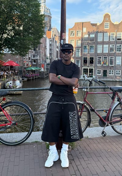 Pantalones cortos oversized negros con el texto "MISBHV", camiseta negra, zapatillas blancas y gafas de sol, de pie junto a un canal con bicicletas de fondo.