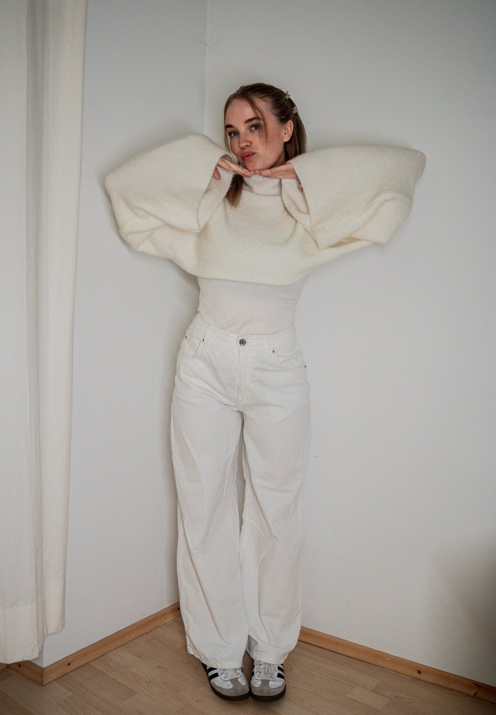 Sweater oversized branco com mangas largas, combinado com jeans brancos de cintura alta. Ténis com detalhes escuros completam o visual.