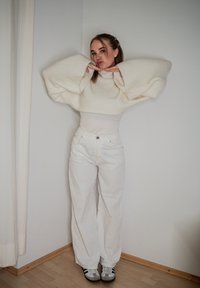 Sweater oversized branco com mangas largas, combinado com jeans brancos de cintura alta. Ténis com detalhes escuros completam o visual.