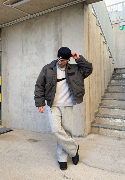 Chaqueta de plumas en verde oscuro con bolsillos de parche sobre una sudadera gris con un logo negro, combinada con unos pantalones de chándal grises y zapatos negros.