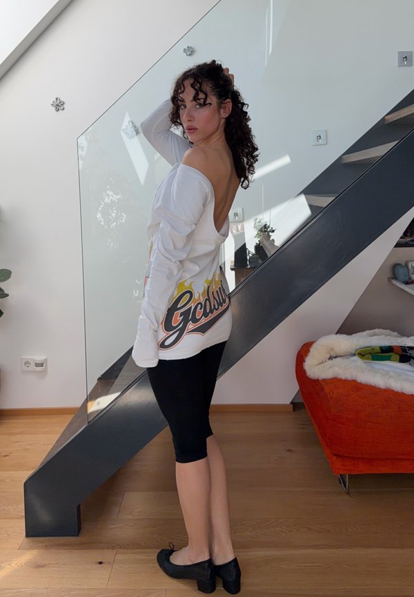 Woman with curly hair wearing a white off-shoulder shirt and black leggings stands by a modern staircase in a minimalist living room.