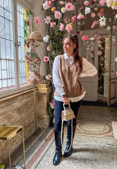 Mujer con chaleco marrón y camisa blanca sosteniendo una pequeña bolsa, de pie cerca de un maniquí floral y flores rosas colgantes en una habitación iluminada por el sol con vidrieras.