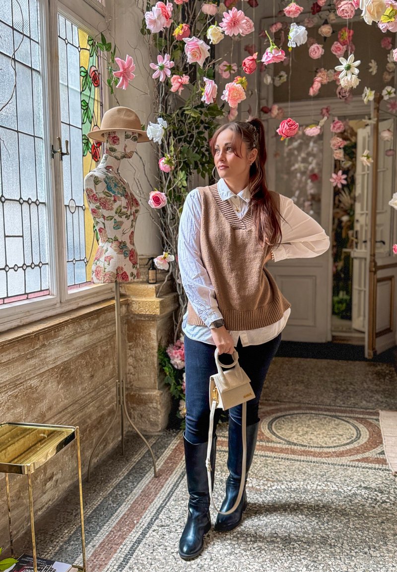 Femme en gilet marron et chemise blanche tenant un petit sac, debout près d'un mannequin floral et de fleurs roses suspendues dans une pièce ensoleillée avec des vitraux.