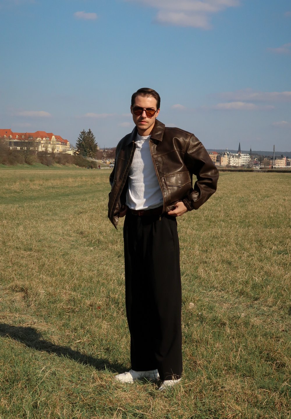 Rjava usnjena jakna s ovratnikom, bela majica, črne široke hlače in bele superge na ozadju travnatega polja.