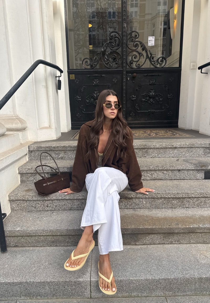 Femeie cu păr lung, ochelari de soare, blazer maro, pantaloni albi și papuci bej, stând pe trepte de piatră lângă o geantă maro în fața unor uși negre ornamentate.