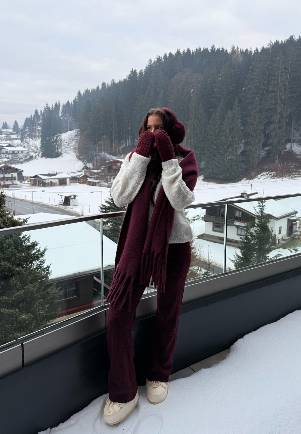 Bufanda de punto granate con flecos, guantes a juego y suéter crema. La persona lleva zapatos crema, de pie sobre la nieve con un bosque de fondo.