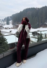 Bufanda de punto granate con flecos, guantes a juego y suéter crema. La persona lleva zapatos crema, de pie sobre la nieve con un bosque de fondo.