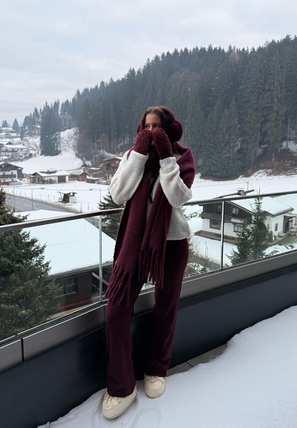 Bufanda de punto granate con flecos, guantes a juego y suéter crema. La persona lleva zapatos crema, de pie sobre la nieve con un bosque de fondo.