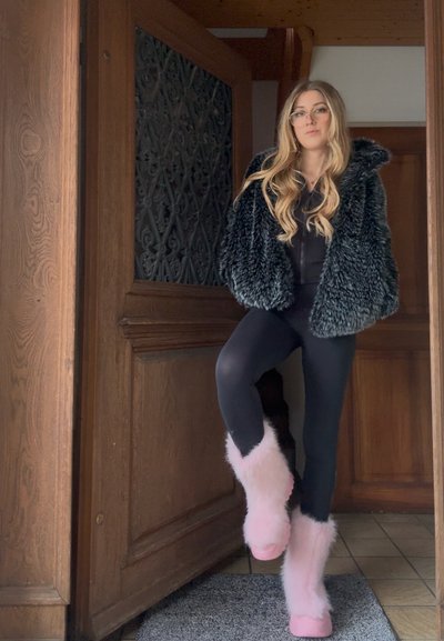 Abrigo negro de piel sintética, leggings negros y botines rosas peludos, con una puerta de madera texturizada y suelo de baldosas de fondo.