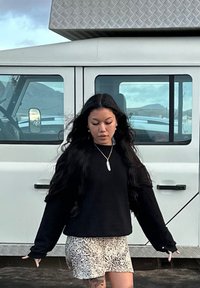 Fekete, hosszú ujjú pulóver, bézs mintás sort, hosszú hajjal és nyaklánccal, egy fehér jármű előtt áll, amelynek fémes, textúrált díszítései vannak.