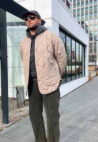 Beige βαμβακερό μπουφάν με καπιτονέ σχέδιο και τσέπη μπροστά, φορεμένο πάνω από μια σκούρα γκρι κουκούλα. Πράσινο ελιές παντελόνι και μαύρα γυαλιά ηλίου ολοκληρώνουν την εμφάνιση.