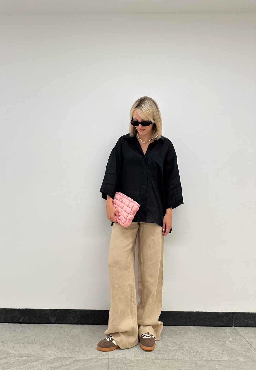 Chemise noire oversize à boutons, pantalon beige à jambes larges, pochette matelassée rose, chaussures marron avec lacets blancs, lunettes de soleil noires, arrière-plan neutre.