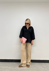 Chemise noire oversize à boutons, pantalon beige à jambes larges, pochette matelassée rose, chaussures marron avec lacets blancs, lunettes de soleil noires, arrière-plan neutre.