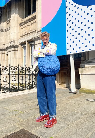 Bolso texturizado azul, camisa floral de gran tamaño, jeans amplios azules y zapatos rojos con cordones azules. El fondo presenta un colorido mural geométrico.