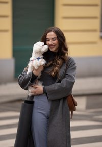 Jeune femme en manteau gris tenant un petit chien blanc près de son visage, souriante, debout près d'une rue avec un bâtiment jaune en arrière-plan.