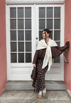 Mujer con falda de lunares, chaqueta marrón, chal blanco y zapatillas, apoyada contra una puerta blanca de paneles de vidrio con una pared rosa.
