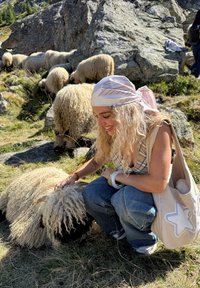 En person kneler ved siden av et fluffy hvitt sau med svart ansikt. Personen har på seg et mønstret hodeplagg og en lys farge på vesken.