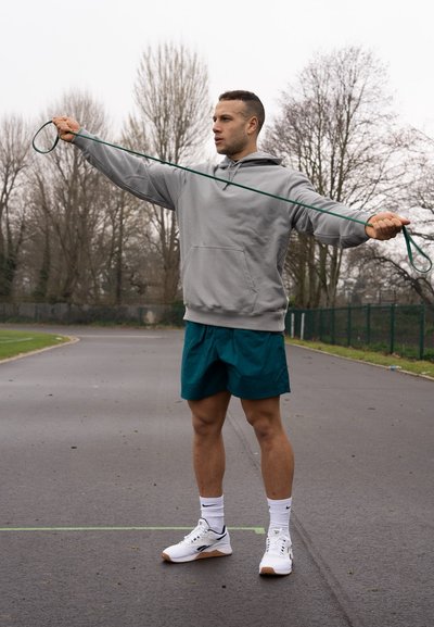 Atleta estirando una banda de resistencia verde, vistiendo una sudadera ligera gris y shorts de color verde azulado, con zapatillas blancas sobre una superficie pavimentada al aire libre.