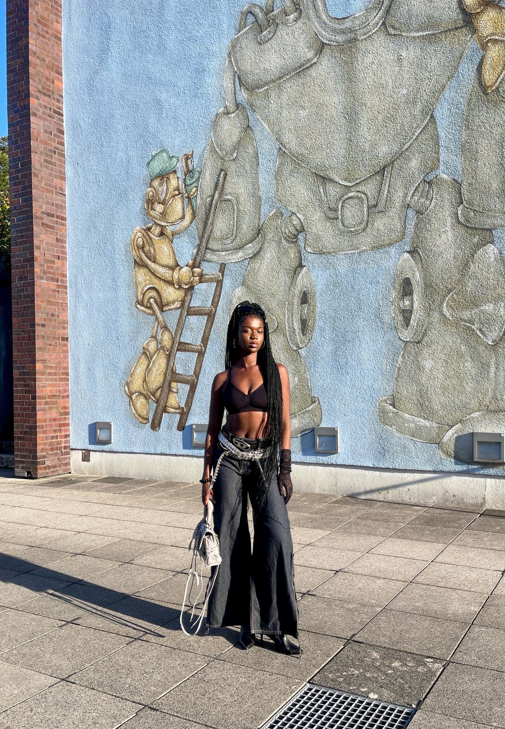 Svart cropped topp, svarte vide jeans og hansker; holder en lys grå veske. Veggmaleri av robotfigurer på en blå vegg i bakgrunnen.