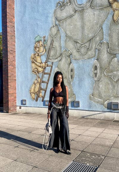 Top corto negro, jeans negros de pierna ancha y guantes; sosteniendo un bolso gris claro. Mural de figuras robóticas en una pared azul de fondo.