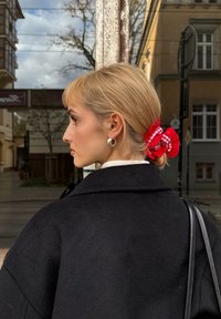 Schwarzer Wollmantel mit hohem Kragen, silbernen Creolen und einem rot-weiß karierten Haargummi, das blonde Haare zurückhält. In der Nähe befindet sich eine reflektierende Oberfläche.