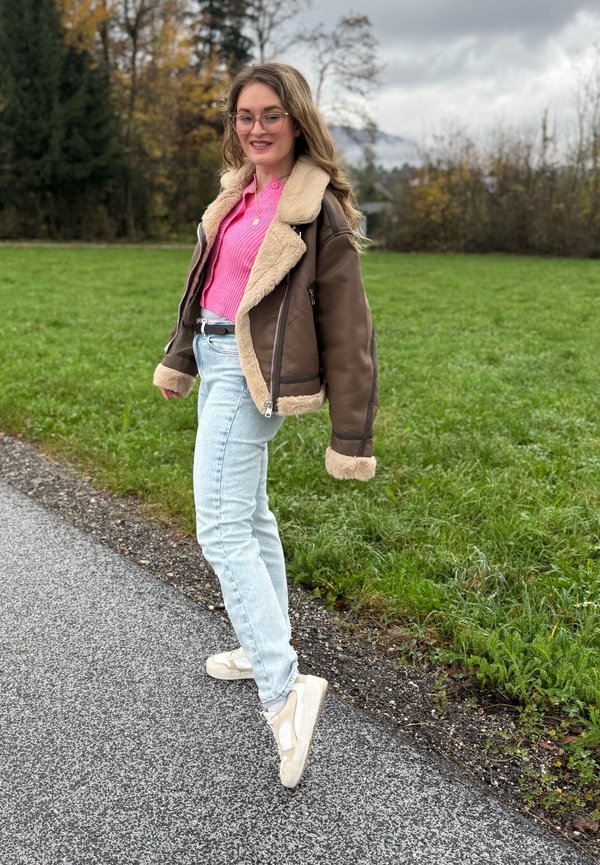 Brown sheepskin jacket with a pale pink top, light blue jeans, and beige trainers. Background features grass and trees under a cloudy sky.
