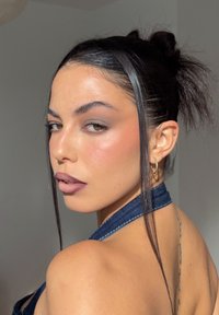 Modelo con cabello largo, liso y oscuro, vistiendo un top de mezclilla con cuello halter. Presenta un maquillaje natural con tonos rosados sutiles y lleva pendientes de aro.