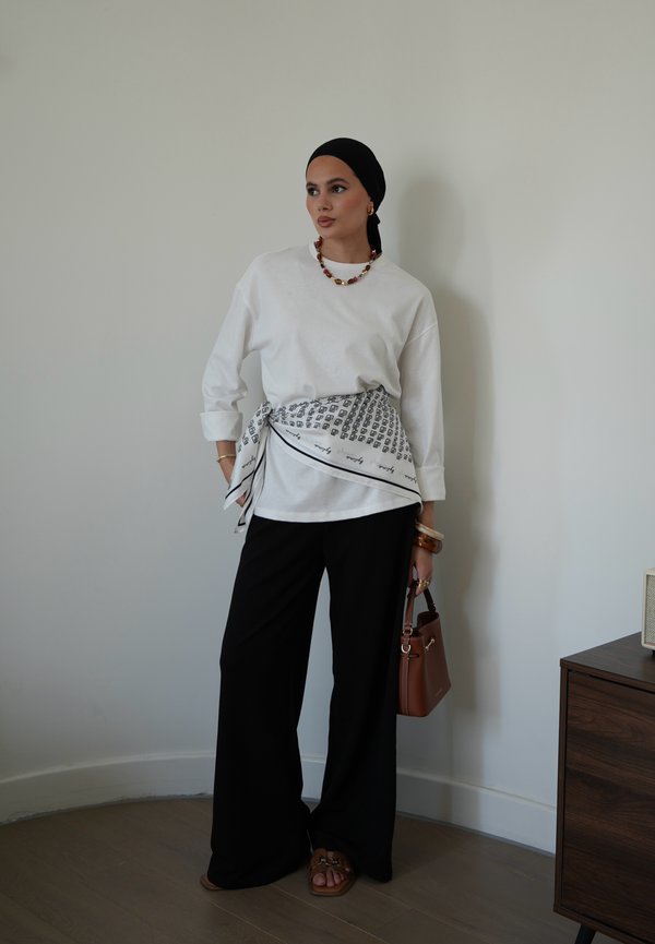 Mujer que lleva una camisa blanca de manga larga, pantalones negros, pañuelo negro en la cabeza, pañuelo estampado atado a la cintura, llevando un bolso marrón en interiores.