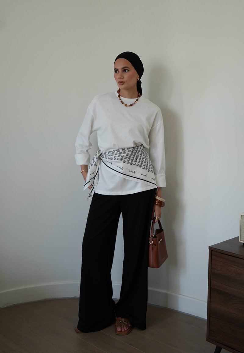 Femme portant une chemise blanche à manches longues, pantalon noir, foulard noir sur la tête, foulard à motifs noué à la taille, portant un sac à main marron à l'intérieur.