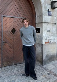 Pull gris en maille avec un col rond, associé à un pantalon en denim noir ample. Debout devant une porte en bois texturée avec un motif en losange.