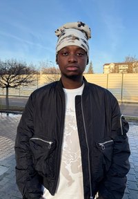 Fekete bomberdzseki cipzáras zsebekkel, fehér grafikás póló fölött, amely egy illusztrációt ábrázol. Világos színű mintás sapka a fején.