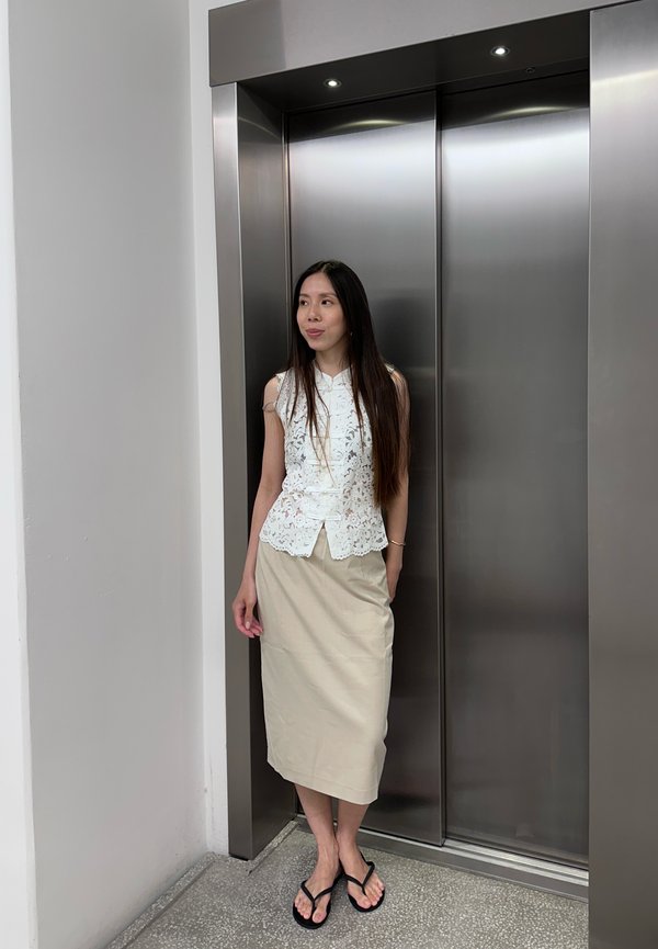 Femme aux longs cheveux portant un top en dentelle blanc et une jupe beige, debout devant des portes d'ascenseur fermées en acier inoxydable.