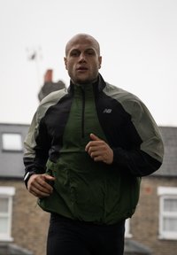 Giacca da corsa leggera verde e nera con collo alto, cerniera frontale ed orlo elastico. Il modello sta facendo jogging all'aperto.