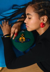 Femme aux cheveux tressés portant des boucles d'oreilles rouges en forme de créoles, reposant les bras sur un coussin bleu, tenant un sac vert avec un emblème de couronne jaune et bleue.