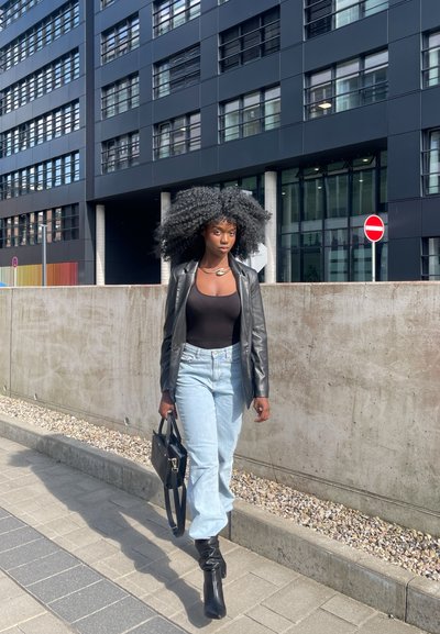 Blazer de cuero negro sobre una blusa negra ajustada, jeans de cintura alta azul claro, botas negras hasta la rodilla, llevando un bolso negro, en un entorno urbano.
