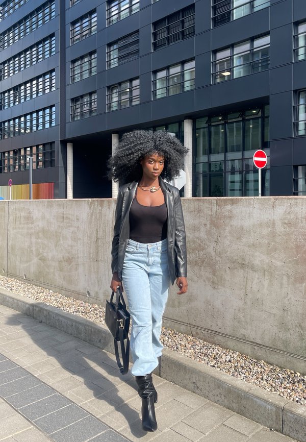 Schwarze Lederblazer über einem figurbetonten schwarzen Oberteil, hellblaue, hochgeschnittene Jeans, kniehohe schwarze Stiefel, eine schwarze Handtasche tragend, städtische Umgebung.