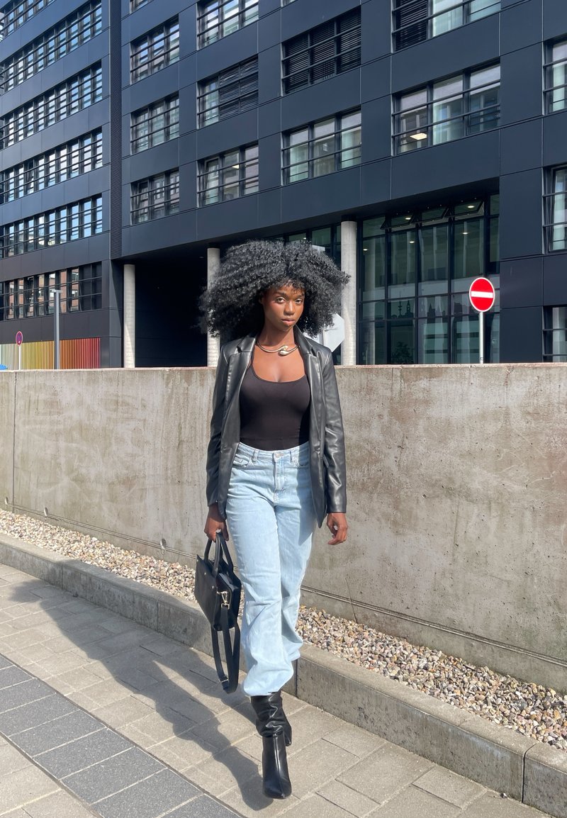 Blazer de couro preto sobre uma blusa preta ajustada, jeans de cintura alta azul claro, botas pretas até o joelho, carregando uma bolsa preta, em um cenário urbano.