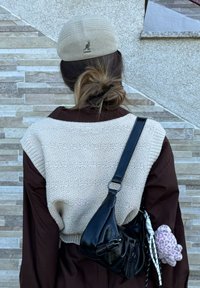 Chaleco de punto crema con hombros anchos, llevado sobre una camisa marrón. Un bolso bandolera negro y un pequeño muñeco de peluche están sujetos a un lado.
