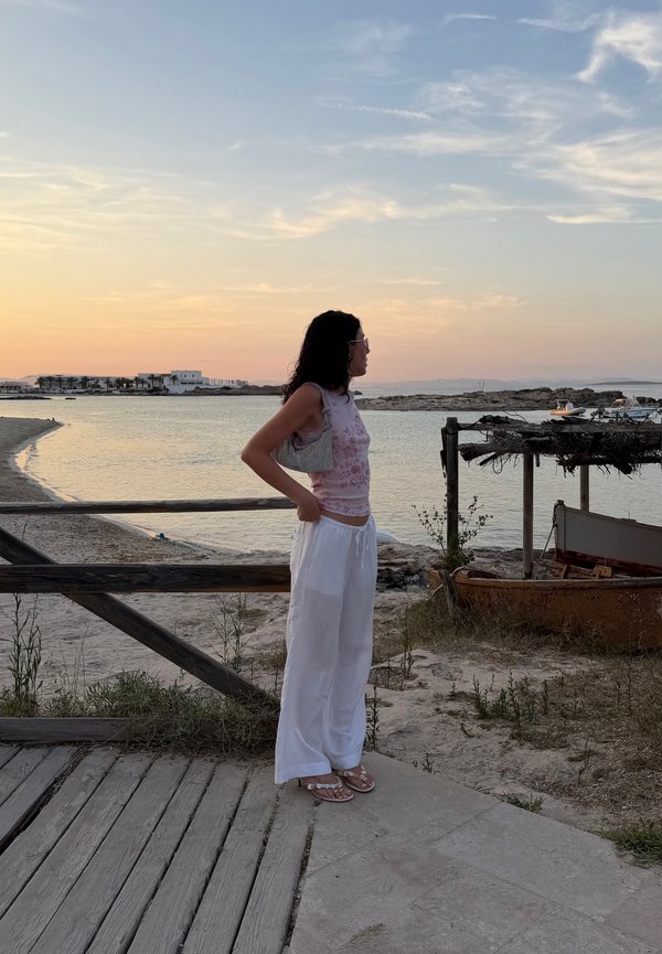 Donna con pantaloni bianchi e top rosa in piedi su una piattaforma di legno vicino all'acqua al tramonto, rivolta verso destra con le mani sui fianchi.