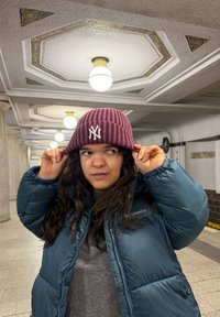 Vinrød ribstrikket hue med hvidt logo foran, båret sammen med en teal farvet dunjakke, i et veloplyst indendørs miljø.