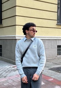 Grijze sweatshirt met een rond logo op de borst, gecombineerd met een lichtblauw overhemd met kraag en donkere broeken. Accessoires zijn onder andere een zonnebril en ringen.