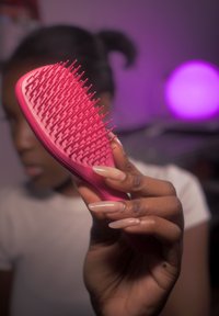 Main tenant une brosse à cheveux rose avec de longs ongles, personne floue aux cheveux attachés et lumière violette en arrière-plan.