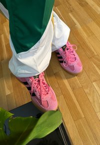 Ροζ σουέτ αθλητικά παπούτσια με μαύρες ρίγες, καουτσούκ σόλα, λεπτομερείς κορδόνες, συνδυασμένα με πράσινα και λευκά παντελόνια προπόνησης και μια δόση από ένα πράσινο φύλλο.