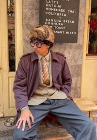 Καφέ πα trench coat πάνω από πουκάμισο με καρό σχέδιο και γραβάτα, γκρι παντελόνι πλατύς καβάλος, φουντωτό καπέλο, μεγάλα γυαλιά και πολλές βέρες στα χέρια.