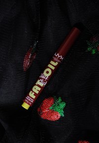 Baume à lèvres NYX Fat Oil dans un tube rouge avec des accents jaunes, présentant une finition brillante. Fond en tissu noir et fraises brodées.