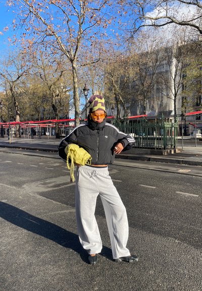 Persona con chaqueta acolchada negra, pantalones grises, gorro estampado, gafas de sol naranjas y zapatos negros con punta dividida sosteniendo una bolsa amarilla en una calle de la ciudad.