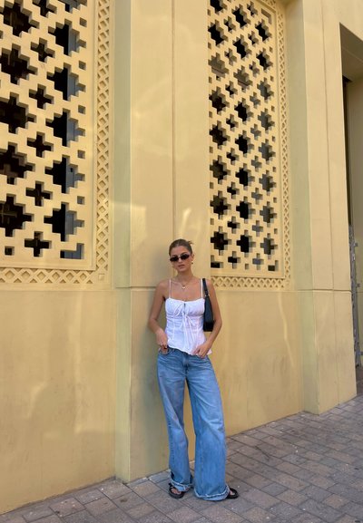 Top corto blanco con detalle de cordón, vaqueros de pierna ancha azul claro, sandalias negras y gafas de sol, posando frente a una pared decorativa amarilla.