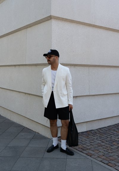 Blazer blanco de lino, pantalones cortos negros, camiseta blanca, gorra negra y bolso negro. Mocasin negro con calcetines blancos. De pie contra una pared de piedra clara.