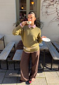 Maglione verde oliva spesso a maglia, camicia rosa chiaro con colletto sotto, pantaloni marroni a gamba larga, che tiene una tazza da tè con piattino.