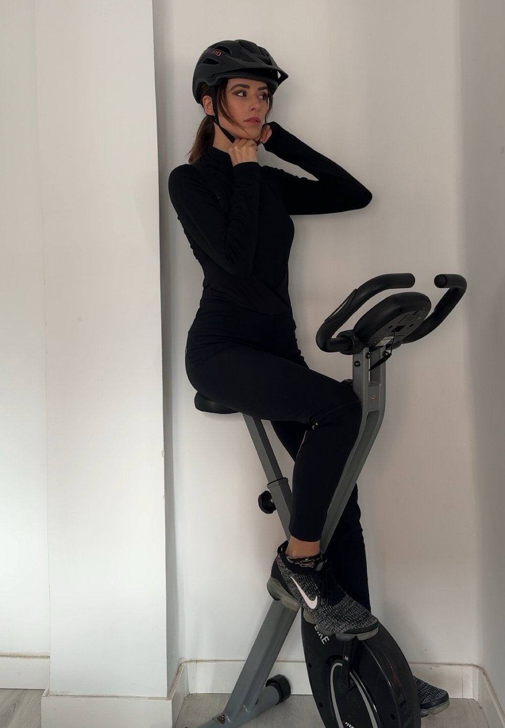 Uma bicicleta de exercício preta com um quadro cinza; uma pessoa usa um capacete preto e um traje de treino ajustado preto, descansando as mãos no pescoço.